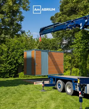 Modular storage container being lifted onto a flatbed trailer by a crane on a grassy area with trees; ABRIUM logo visible.