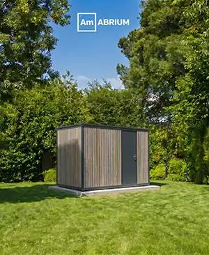 Small wooden garden shed with a gray door on a concrete base in a sunny, green backyard with trees. Logo saying 'AmBRIUM' at the top.