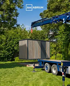 Modular container being hoisted onto a blue trailer by a crane in a grassy yard; AmBRIUM logo at the top.