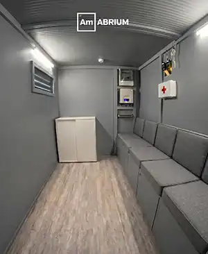 Interior of a small grey mobile clinic or van: bench seating along the right wall, a white cabinet at the far end, and medical equipment mounted on the wall including a first aid kit sign.